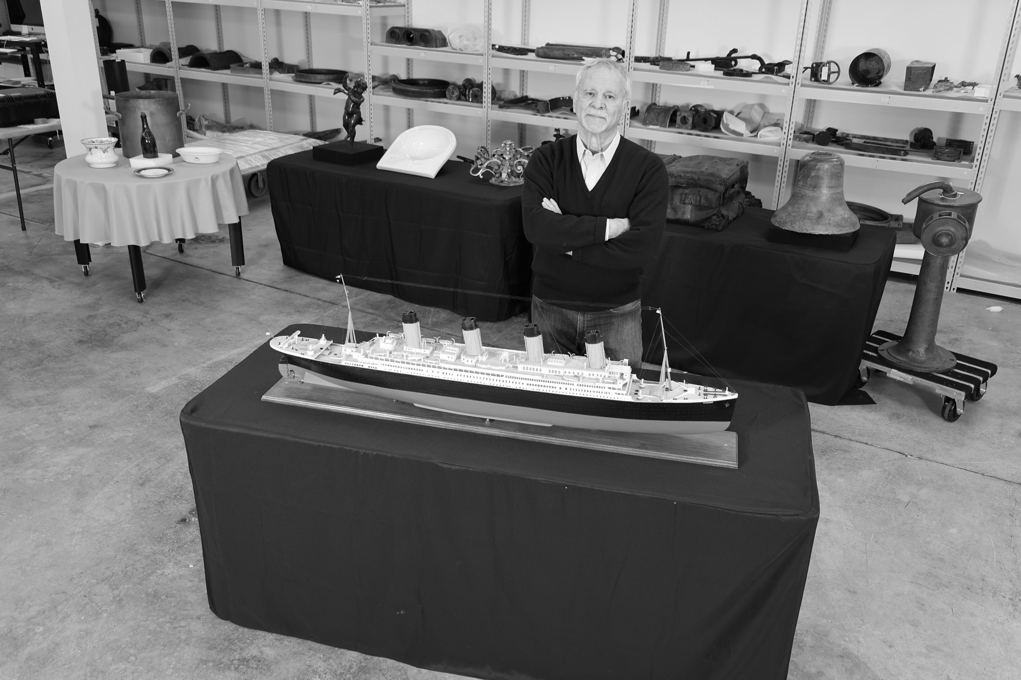 A black and white photo of an older man standing behind a model of the Titanic boat.