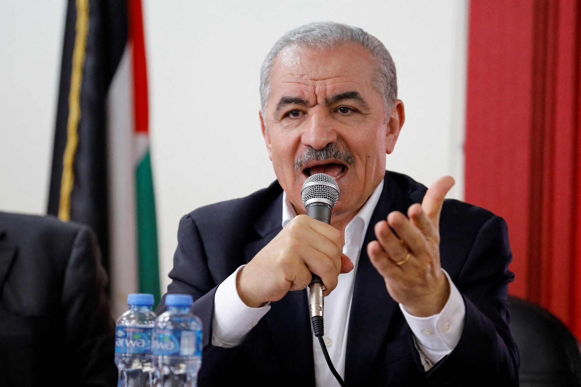 An older Palestinian man with short hair and a neat moustache speaks into a microphone while seated.