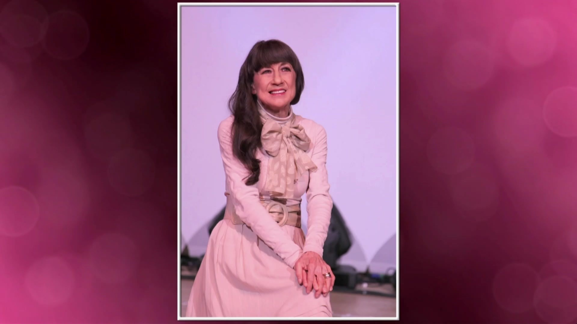 Judith Durham smiles as she sits in a photo against a pink backdrop.
