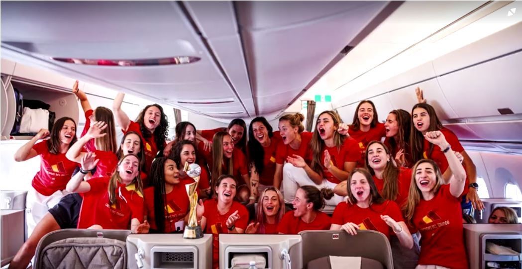 A group of spanish people in red unifroms on a plane posing with trophy