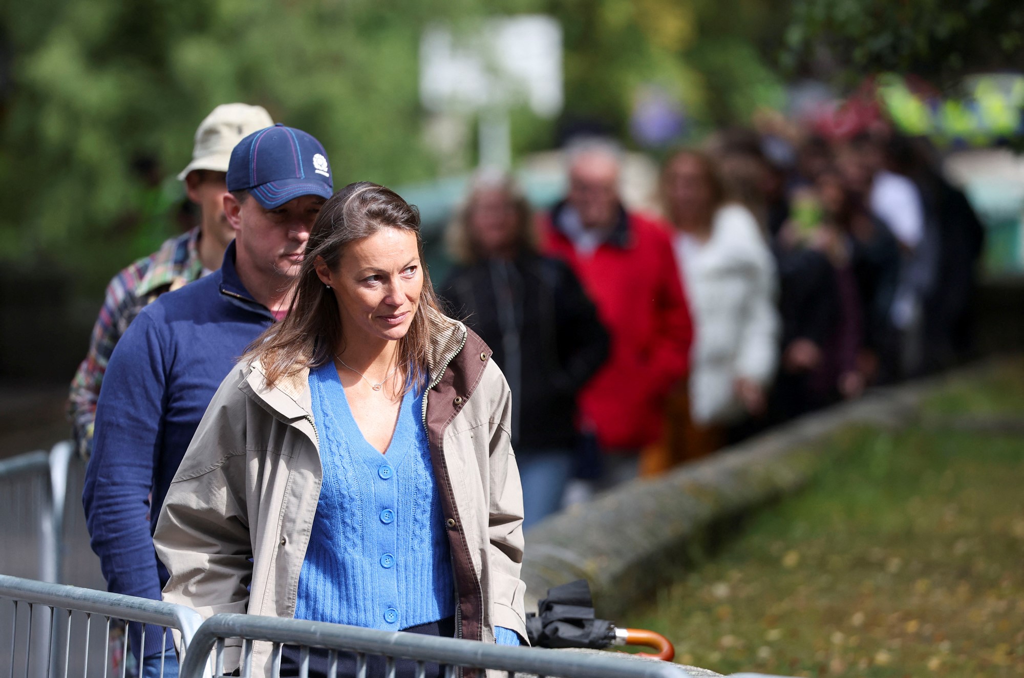 People stand in a line behind a barrier near grass.