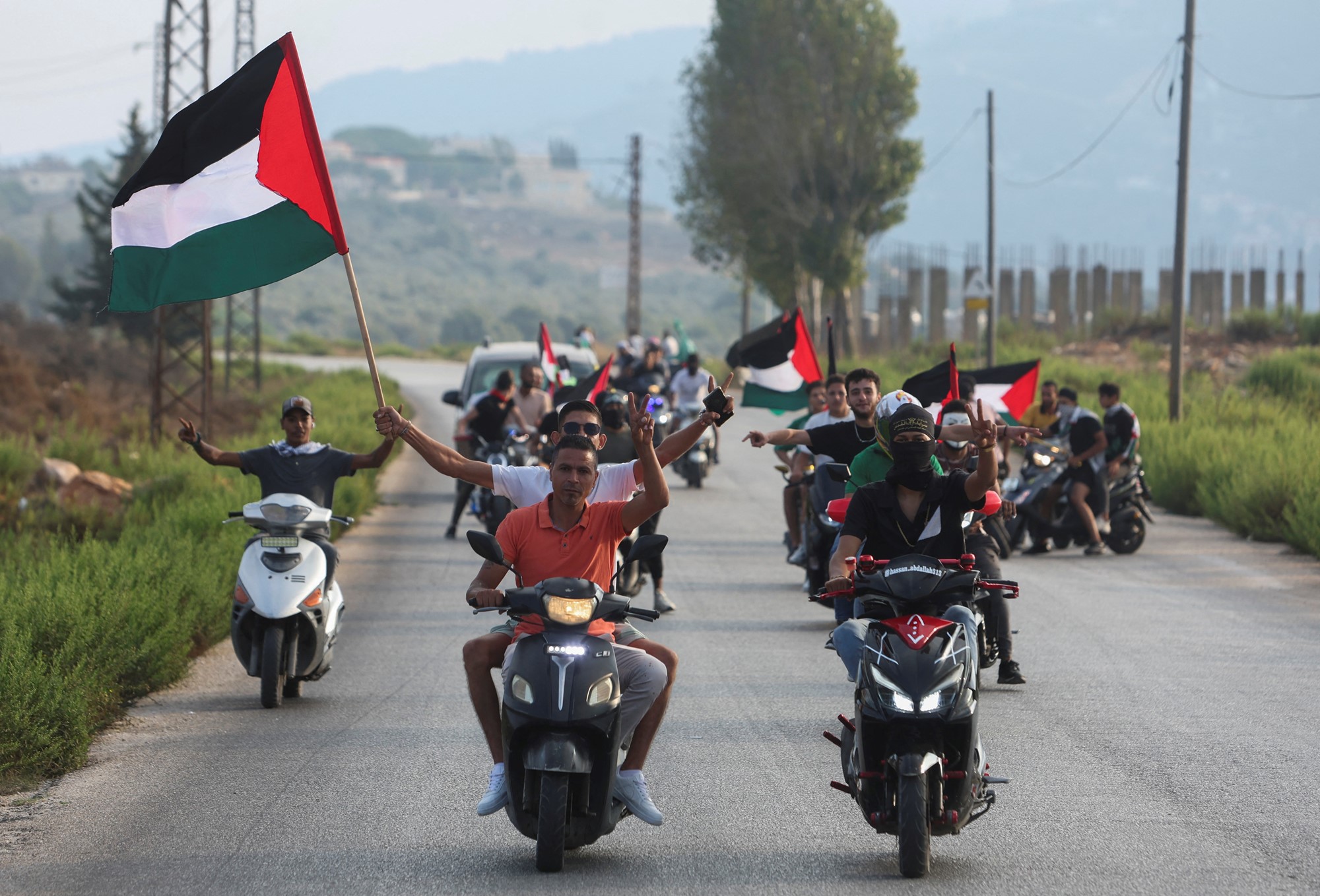 Several men on motorbikes, waving flags and making the peace sign with their hands.