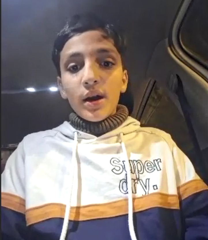 A young boy wearing a hoodie sits inside a car speaking to a camera
