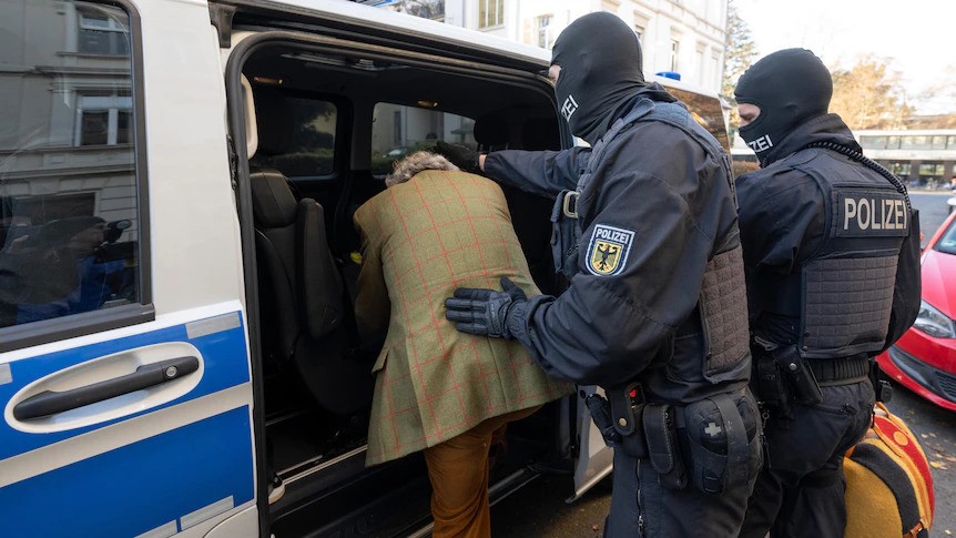 A man in a suit is pushed into a van by two masked police. 