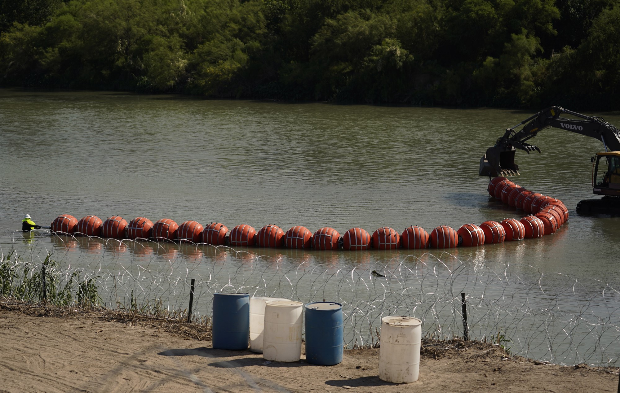 A wide shot of a connected line of plastic buoys in a body of water, being pulled by an excavator