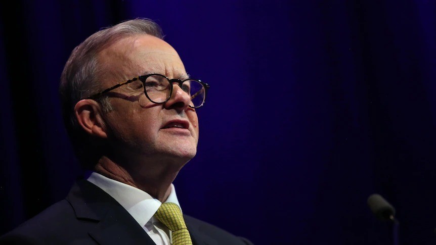 Close up of Anthony Albanese. He wears glasses with a black frame and a suit with a yellow tie.