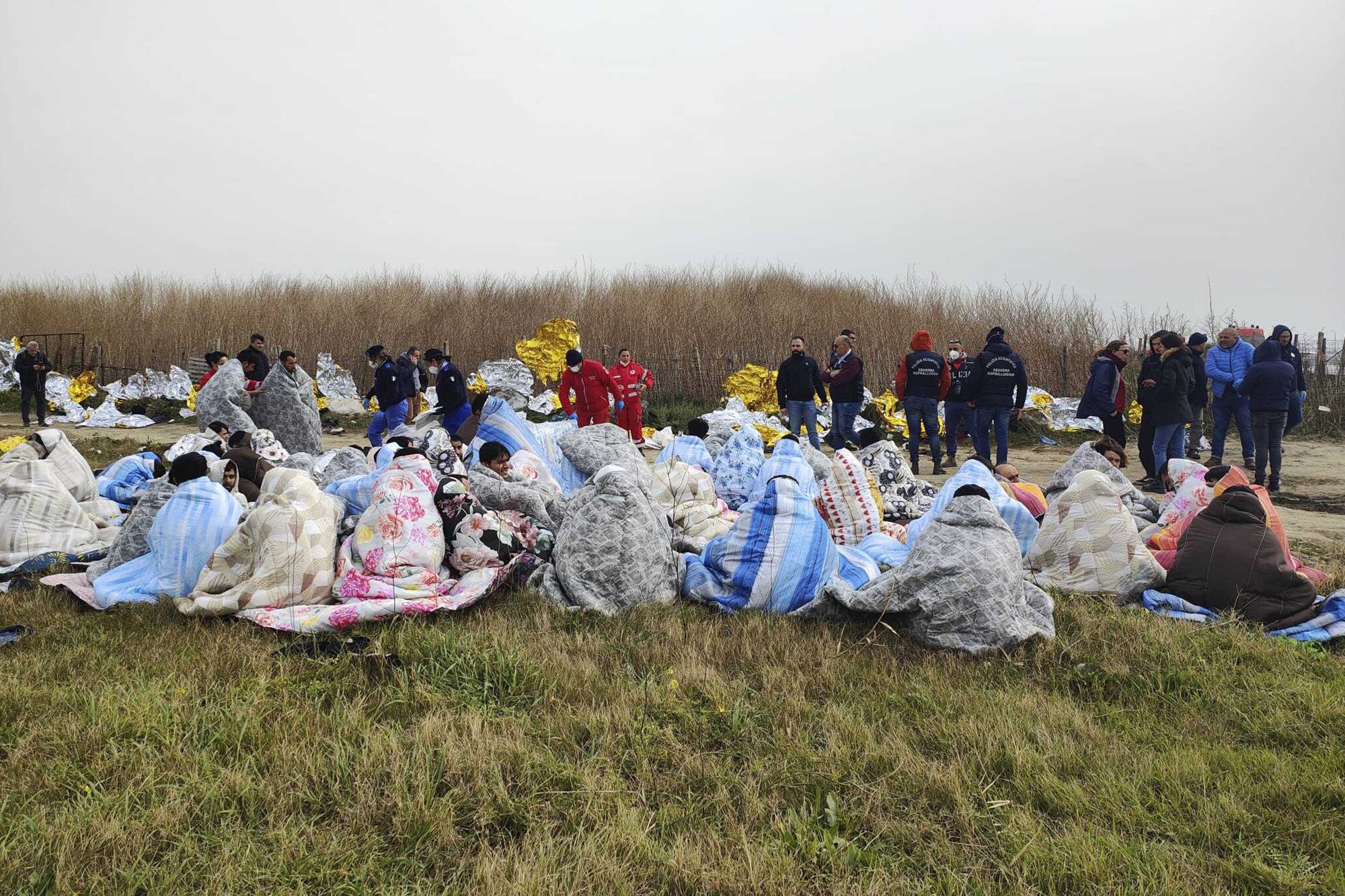 a group of people all wrapped in blankets sit on grass as emergency workers stand with shock blankets in the background. 