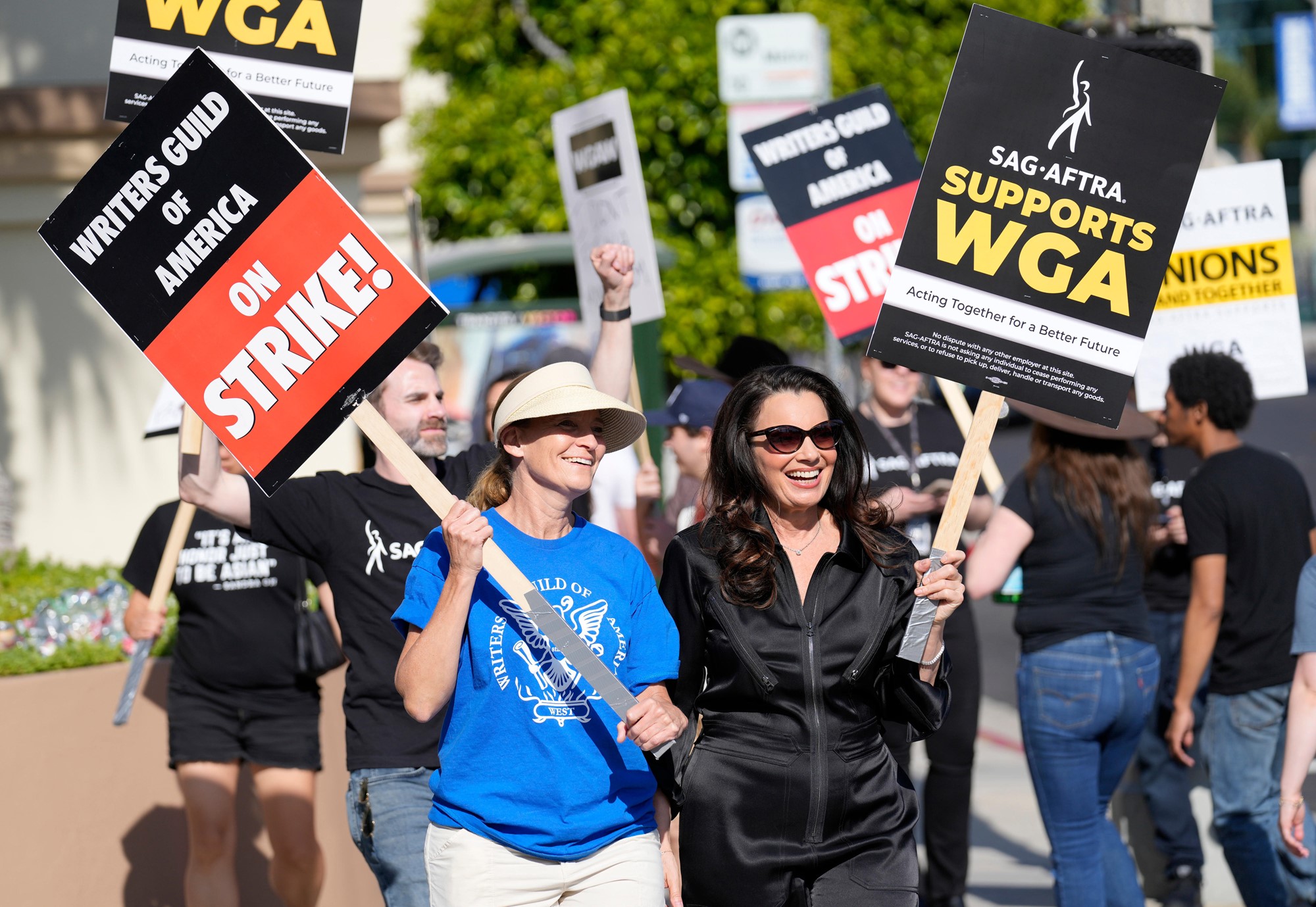 Fran Drescher in black velvet tracksuit and Meredith Stiehm in blue T smile while holding protest signs