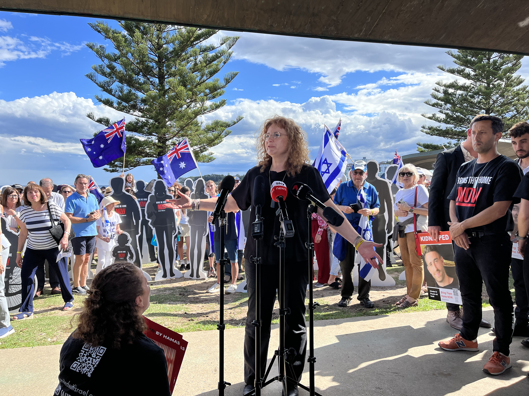 A woman speaking at a beach with a crowd holidng Australian and Israel flags. 