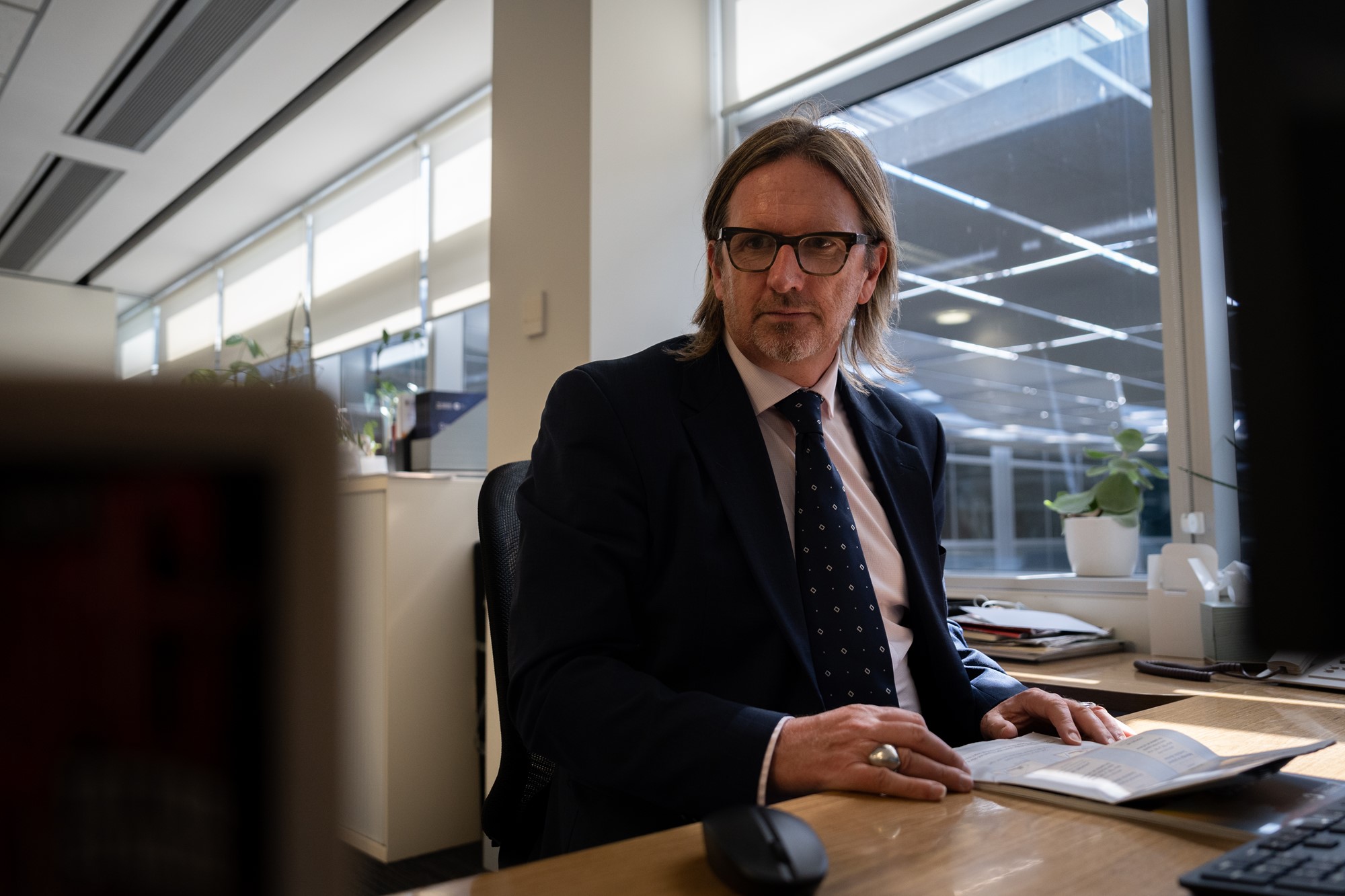 A middle-aged white man in a suit with shoulder-length hair, a beard and glasses.