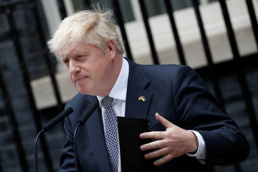 Boris Johnson stands in front of a microphone and gestures with his left hand.
