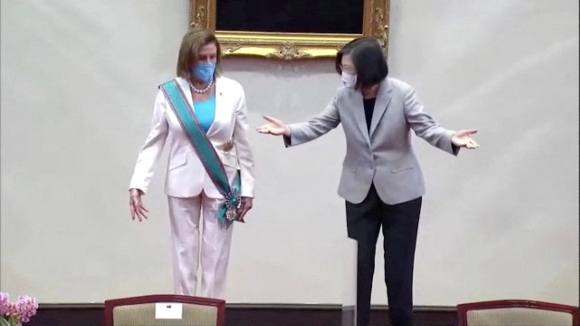 A woman wearing a white suit and aqua cash stands next to a woman wearing a grey blazer gesturing.