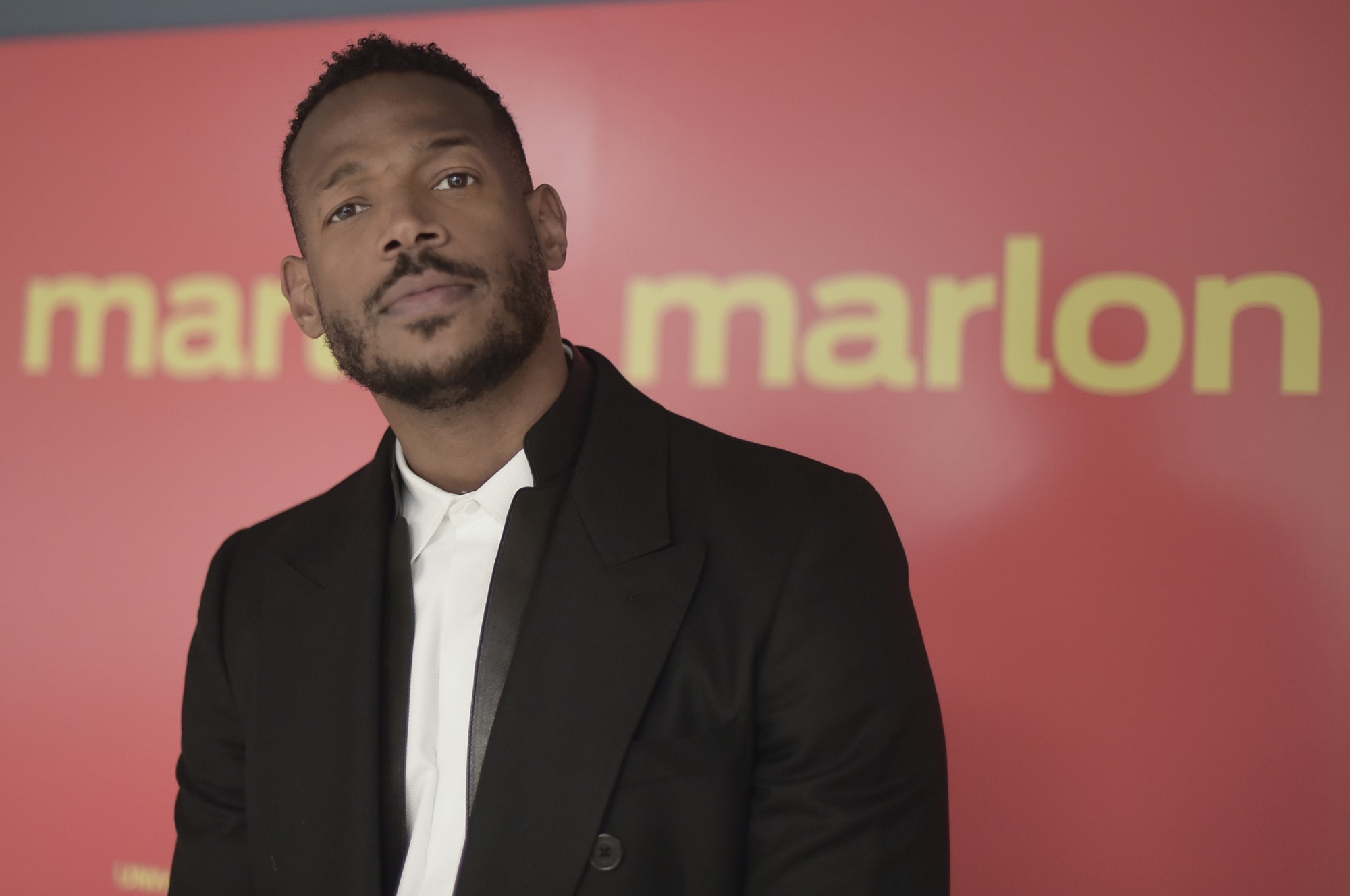 A black man in a suit stands in front of a sign which reads 'Marlon'