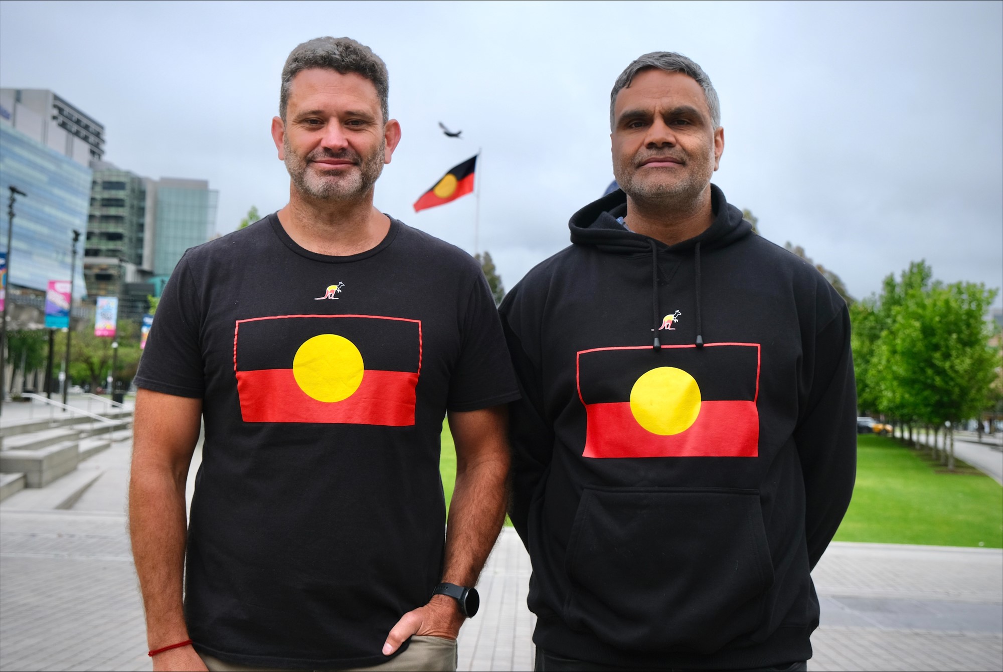 Two men stand wearing Aborignal flag t-shirts.