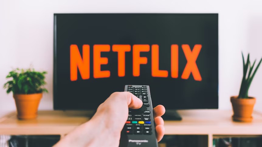 A hand holding a TV remote points at a TV, which is displaying the Netflix logo