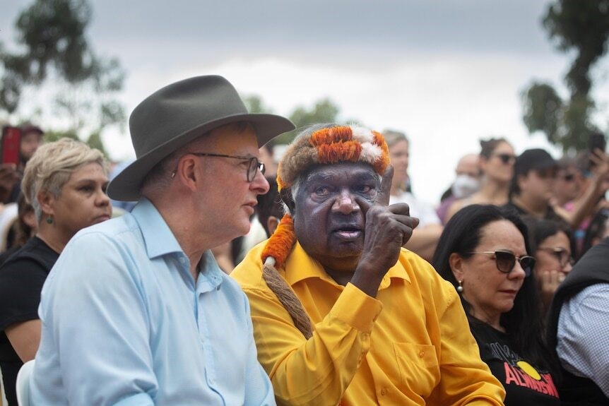 Yunupingu sits next to Anthony Albanese, and holds up a finger while looking at the Prime Minister.