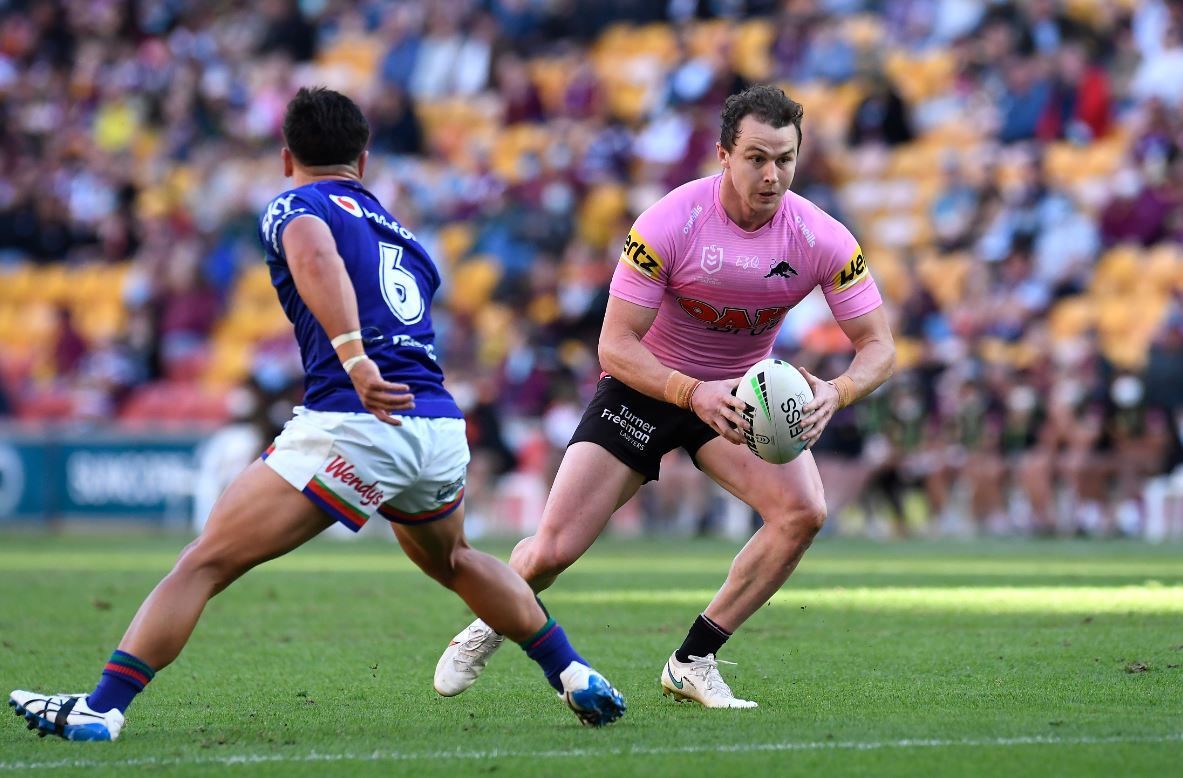 An NRL player in pink, holding the ball in both hands, steps past a player in blue with the number 6 on his back