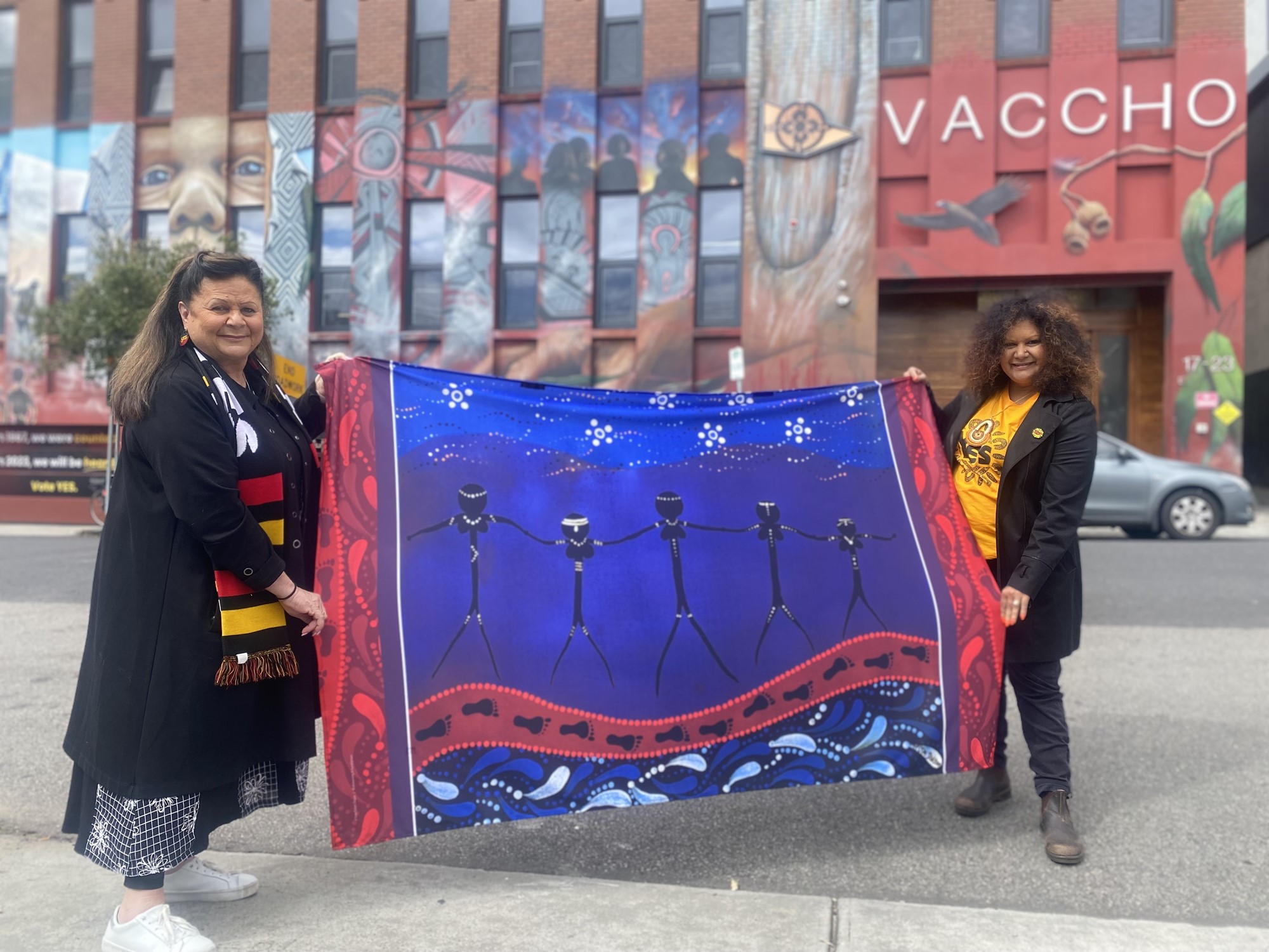 The two women hold up a blue shawl with five figures painted on it.