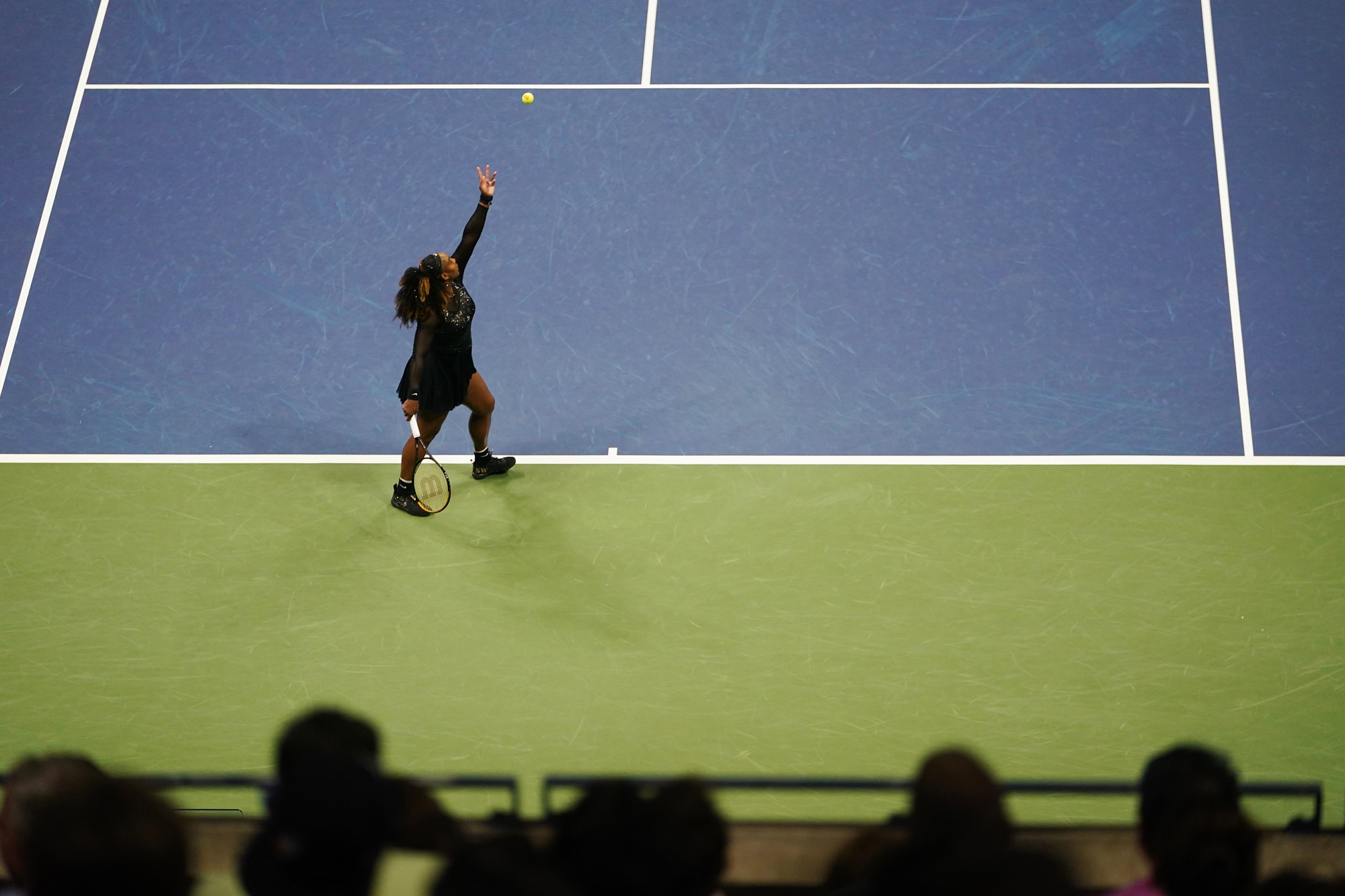 A woman in all black serves a tennis ball at the US Open.