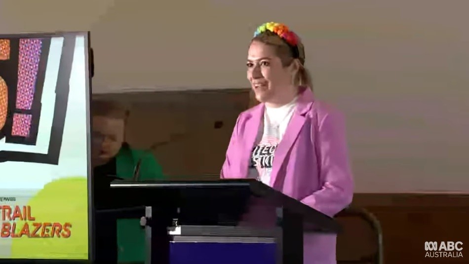 A young woman with a pink power suit and rianbow headband speaks at a podium.