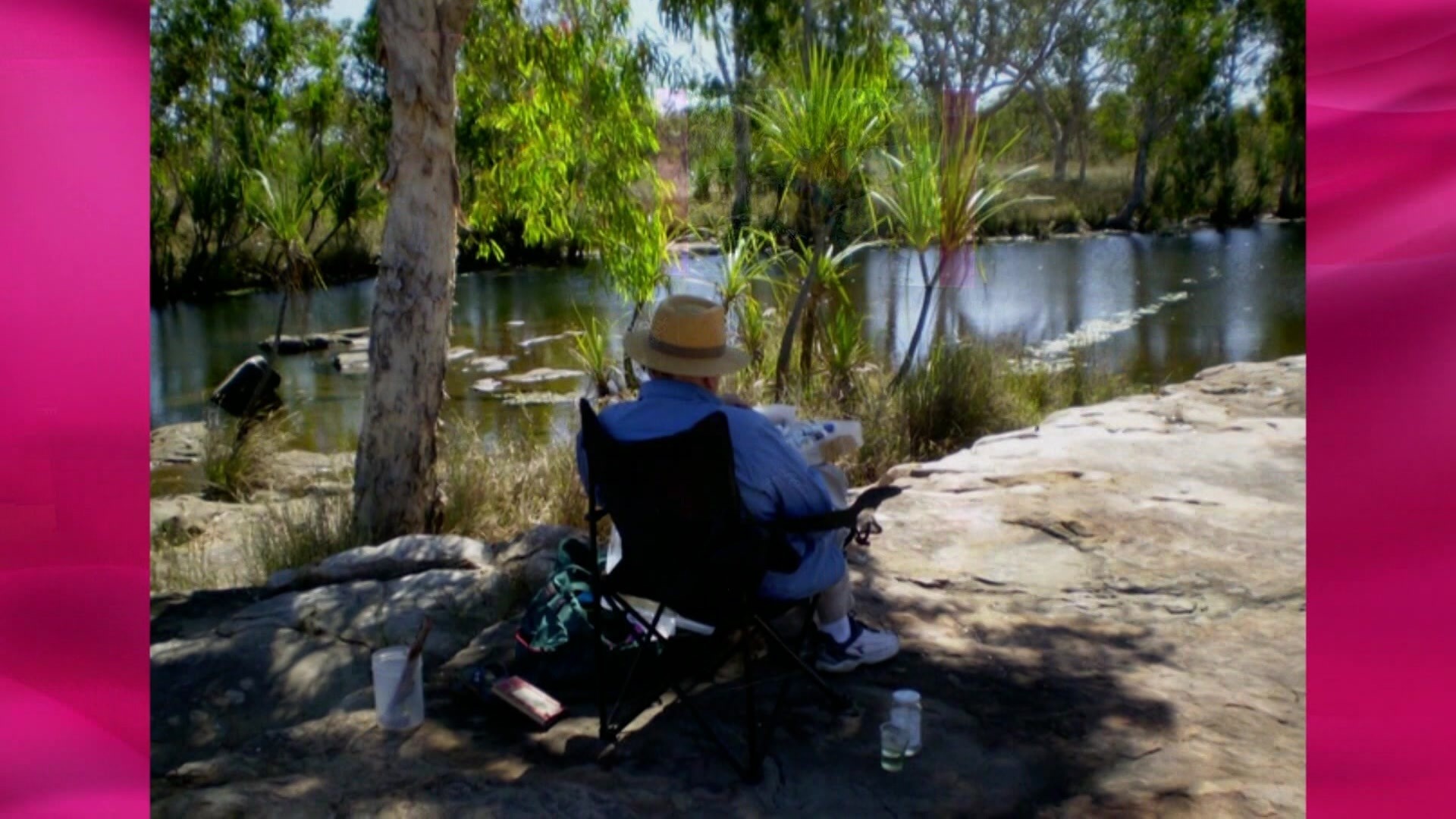 Barry  Humphries on a chair in the bush with his back to the camera.
