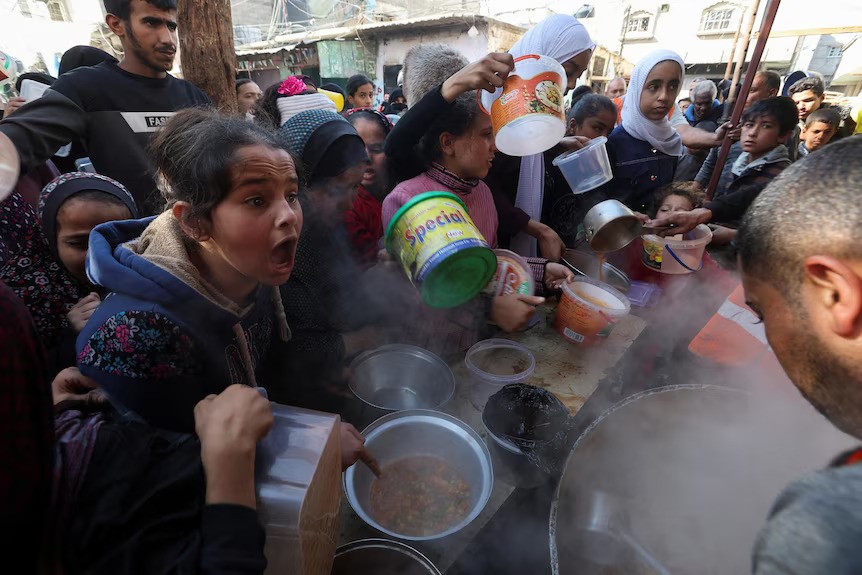 A crowd of people hold out buckets around a man distributing food from a massive steaming pot