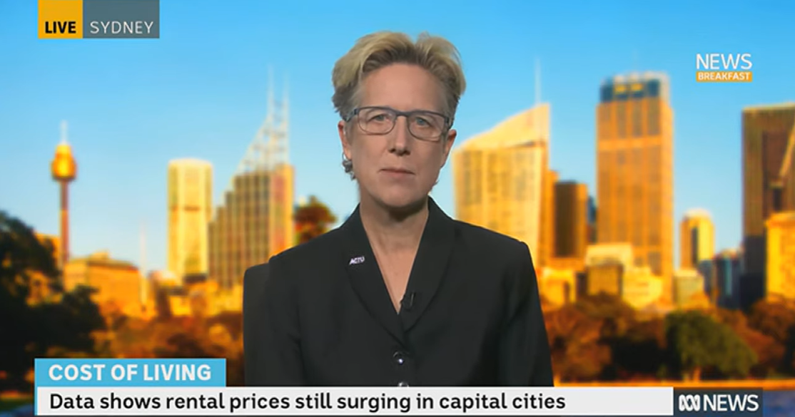A woman with short hair and glasses does a TV interview in front of a Sydney skyline backdrop