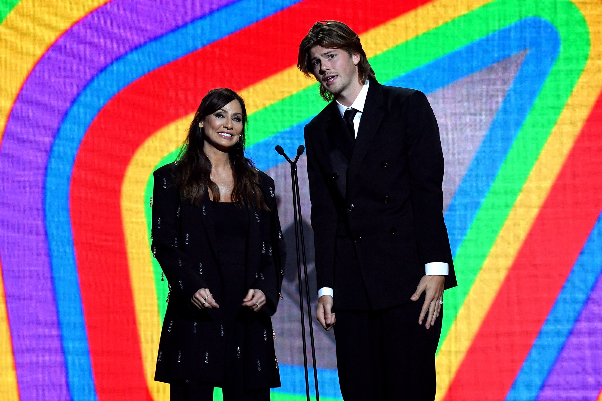 natalie imbruglia and ruel stand side by side on stage in front of a rainbow background presenting an award
