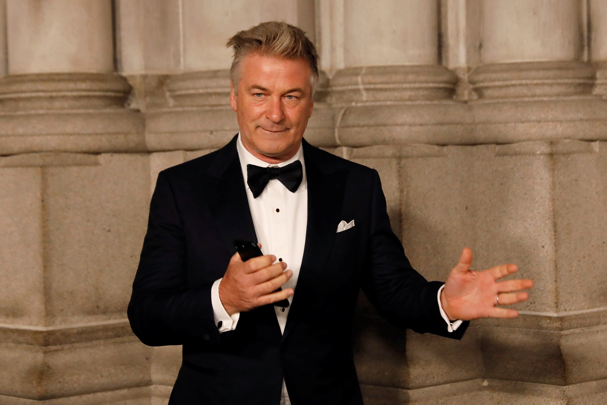 Alec Baldwin wears a suit and gestures with his hands. 