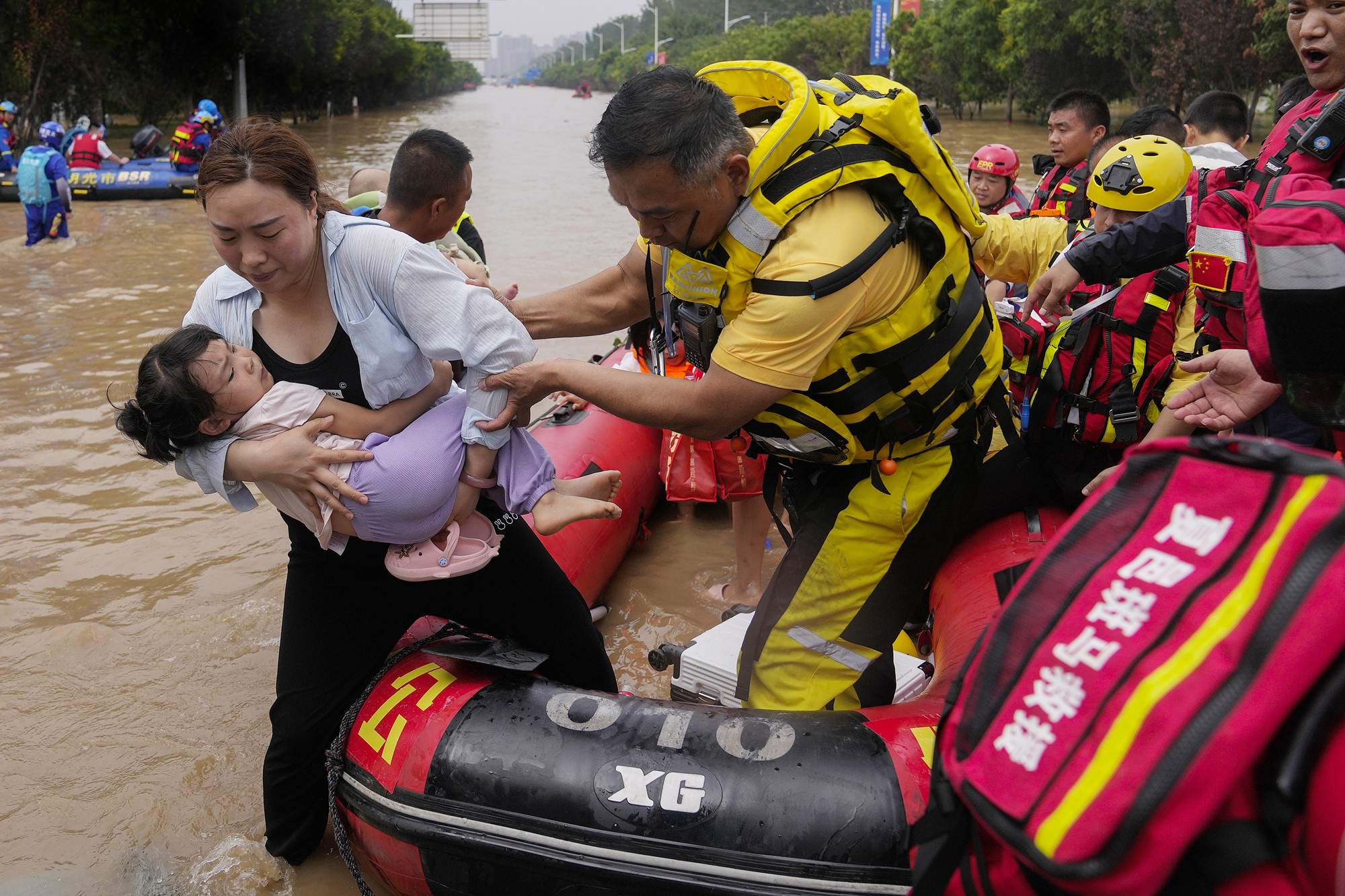 A woman holding a young girl steps out of a small rubber boat, helped by a man, in floodwaters over a road. Others in the boat behind them.