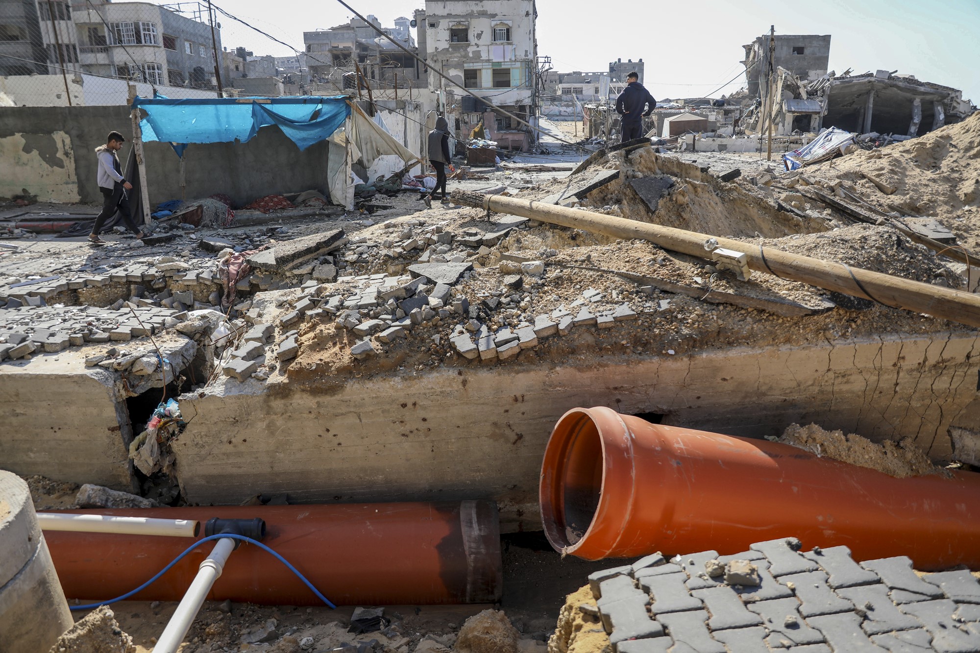 A medium shot of a broken large pipe, lying alongside rubble near people walking and badly damaged buildings in the background